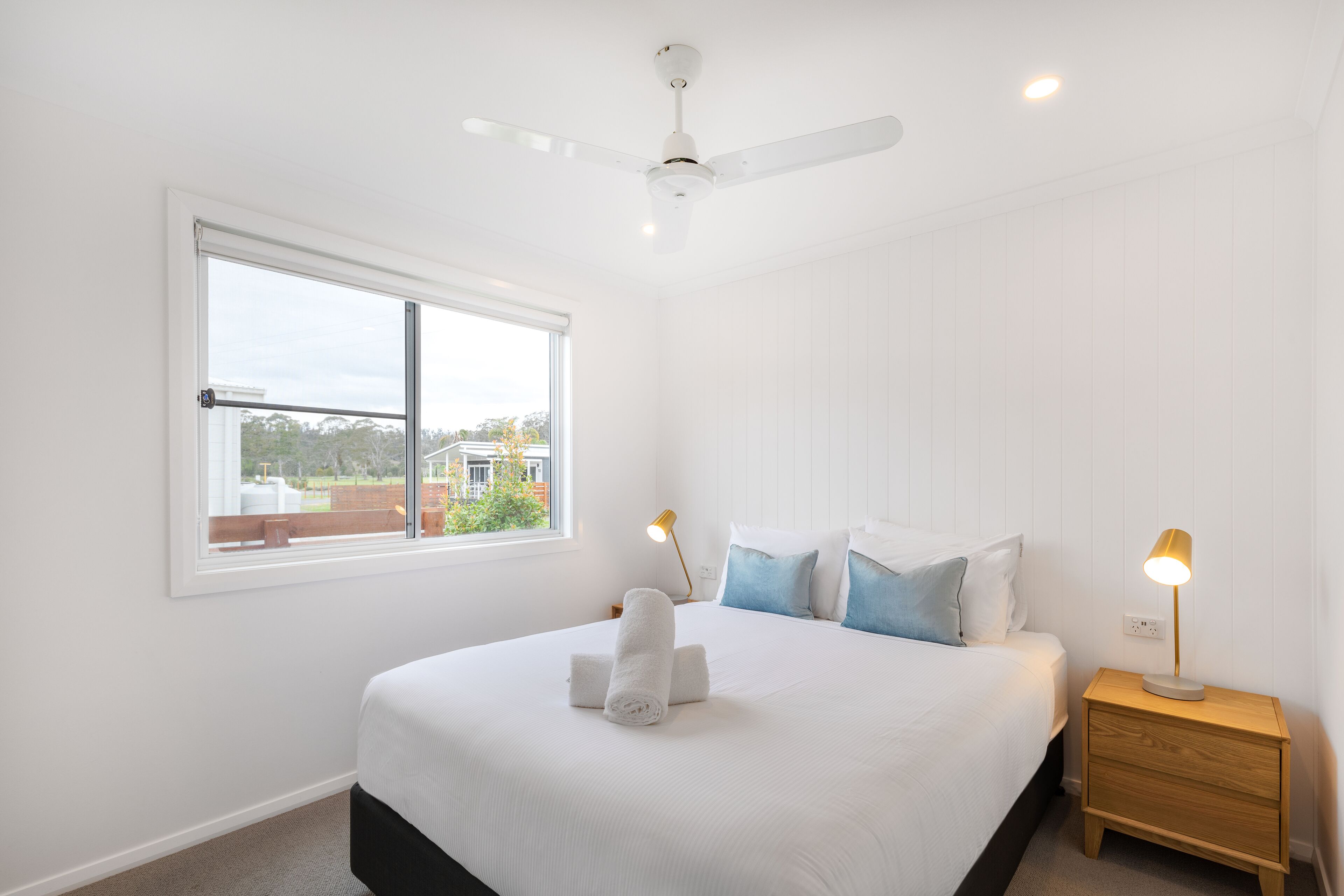 A bedroom with a large bed, a ceiling fan, a nightstand with a lamp, and a window with a view of the outside.