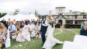 A wedding ceremony outdoors with guests seated on white chairs, a building in the background, and a grassy area.