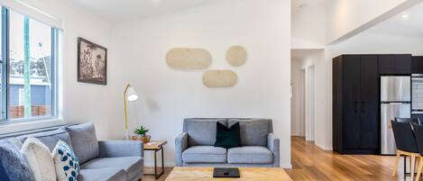 Ein modernes Wohnzimmer mit einem grauen Sofa, einem Holztisch und einer Küchenzeile mit dunklen Schränken und einem Edelstahlkühlschrank.
