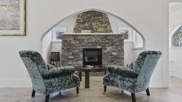 A room with a stone fireplace, two upholstered chairs, a round table, and a framed picture on the wall.