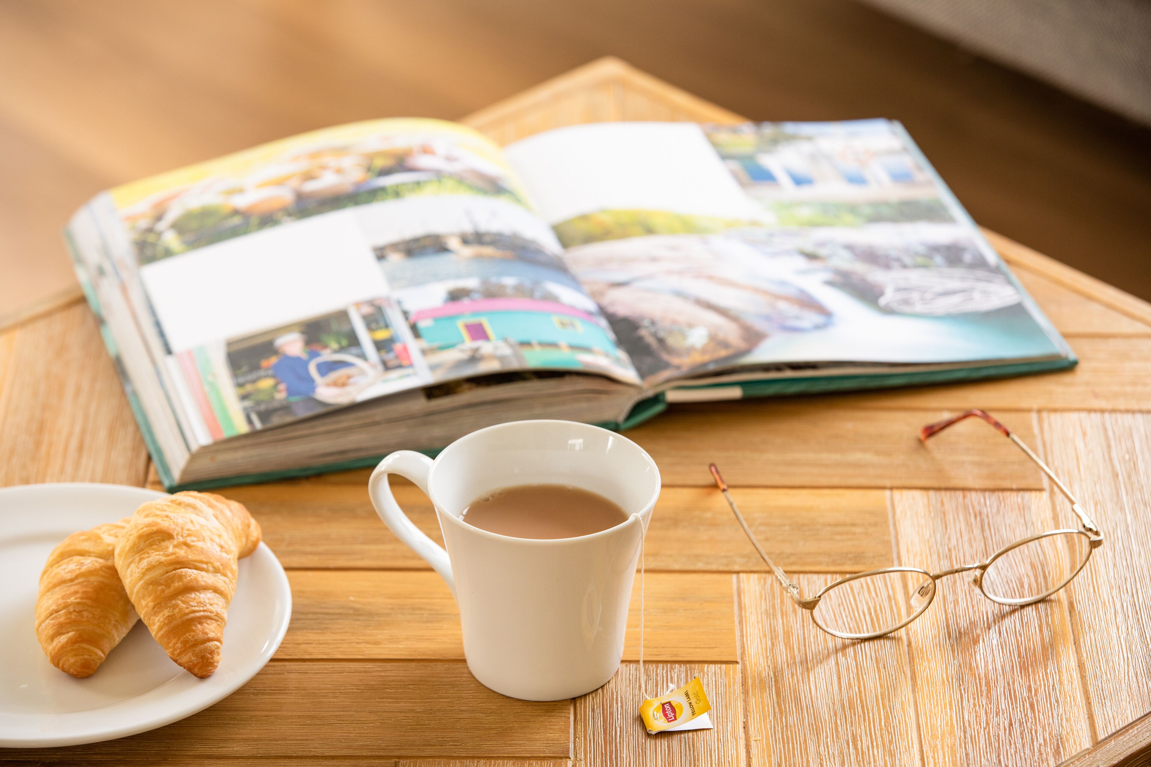 Une table avec une tasse blanche, une paire de lunettes, une assiette contenant des croissants et un magazine ouvert.