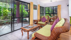 A spacious living area with a large glass door leading to a balcony, wicker furniture, and a ceiling fan. - Dreamcatcher Beachsides Apartments (Port Douglas)
