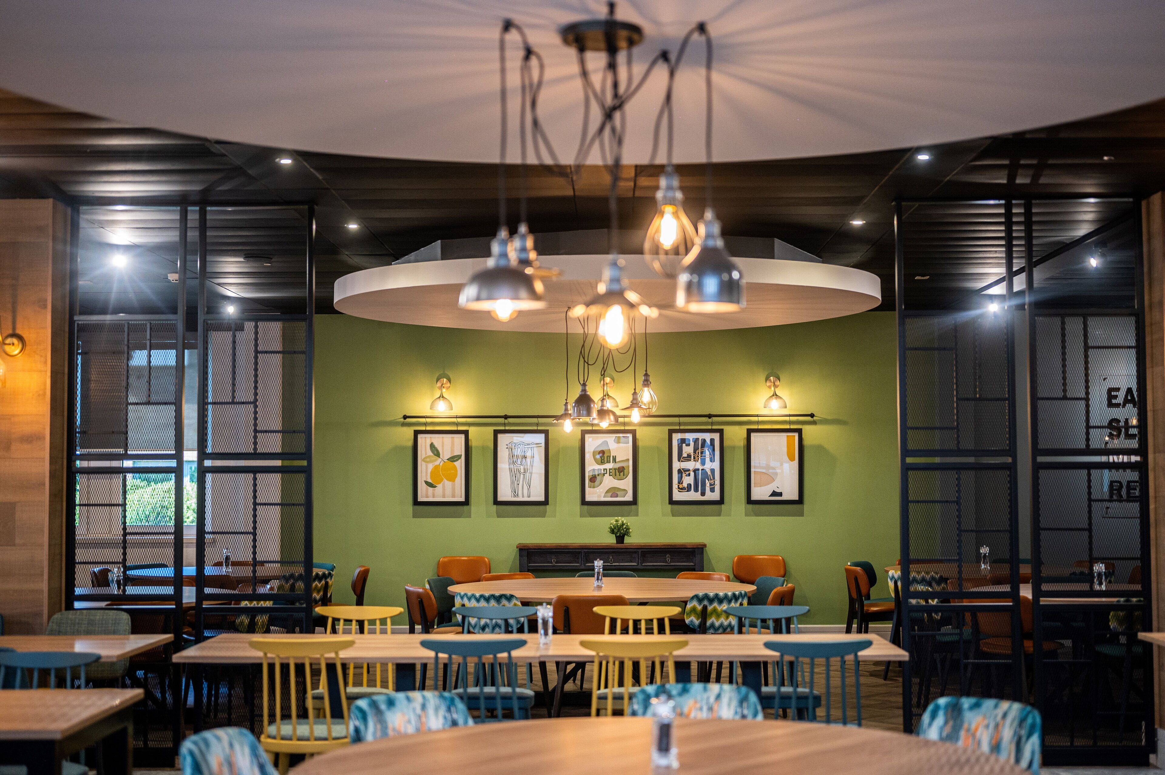 Un restaurant doté de nombreuses tables et chaises, d’un mur végétal agrémenté de tableaux encadrés et de luminaires suspendus.