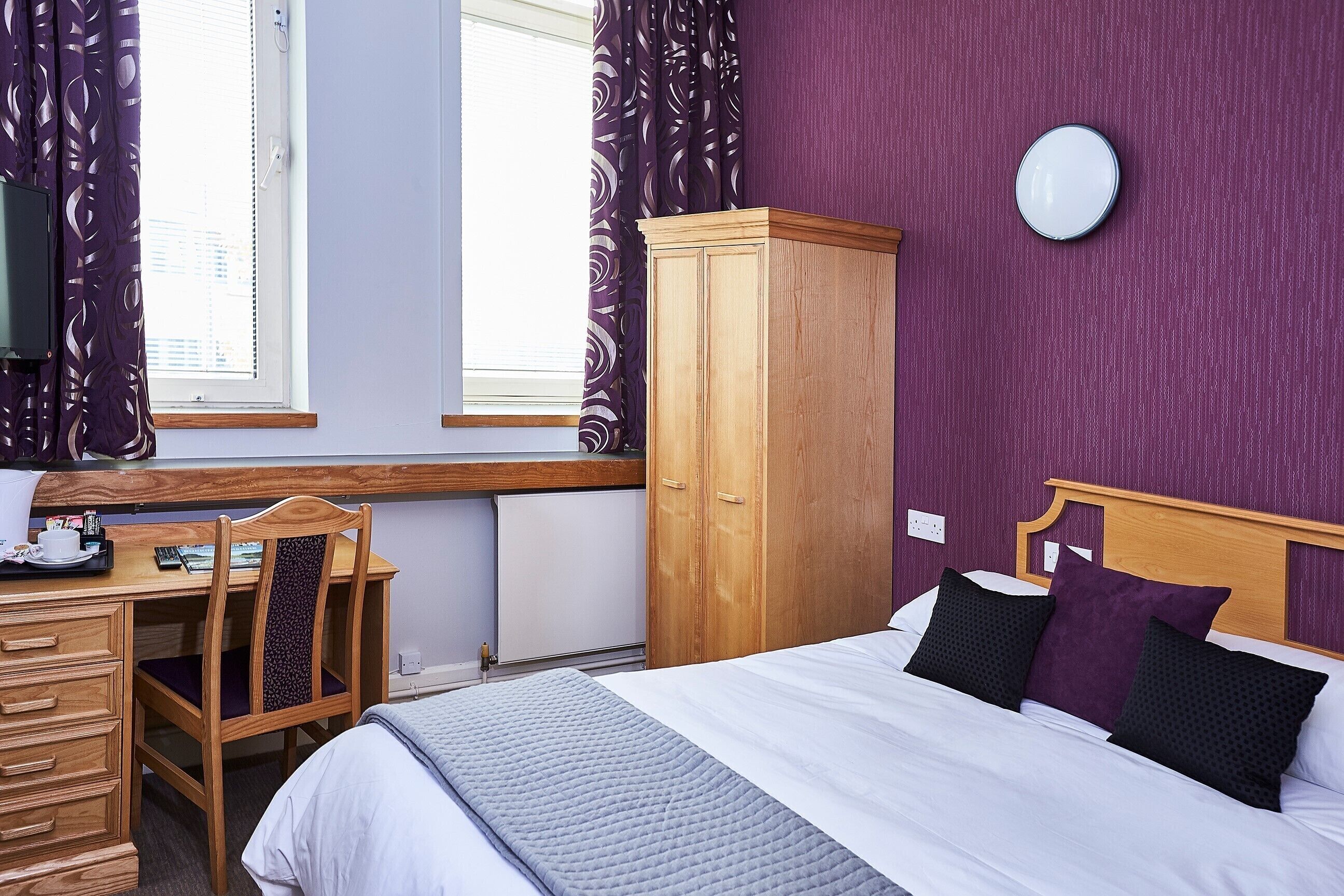 A bedroom with a bed, a wooden desk, a chair, a wardrobe, and a window with curtains.