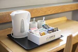 Habitación de hotel con cafetera, dos tazas y una caja de cápsulas de café Clipper sobre una mesa de madera.