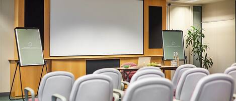 A conference room with a large screen, white chairs, a table with a laptop, and two flip charts.