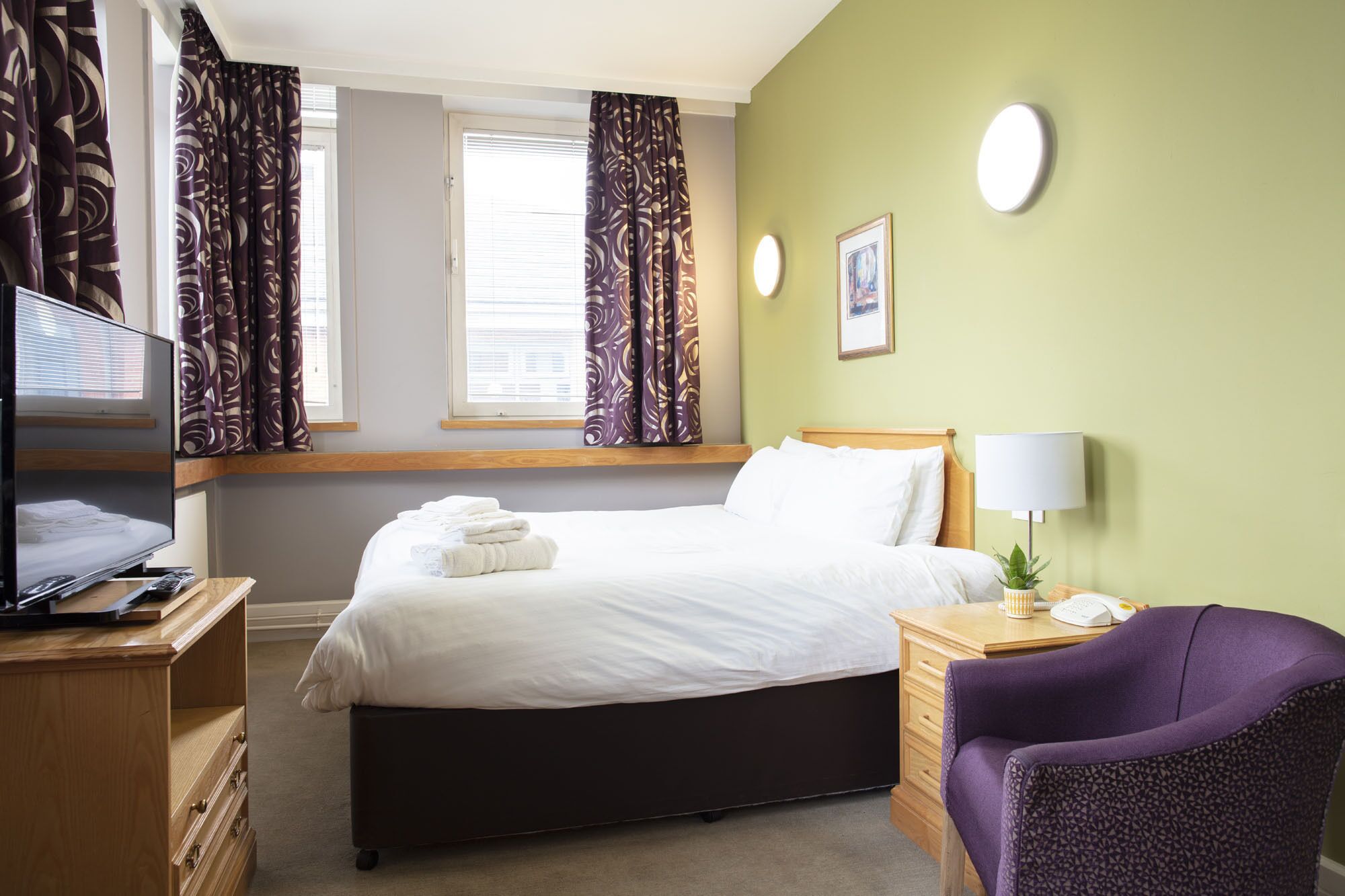A hotel room with a bed, a desk with a lamp, a chair, a television, and a window with curtains.