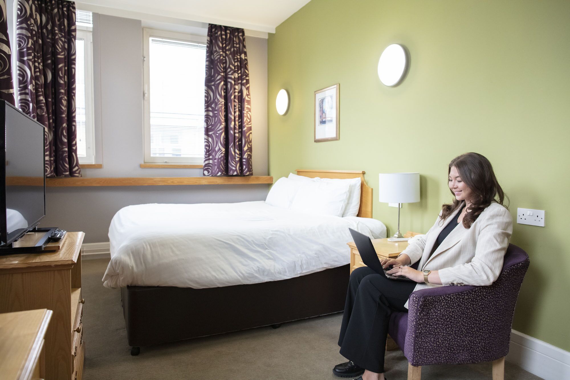Een hotelkamer met een bed, een bureau met een laptop, een stoel en een lamp.
