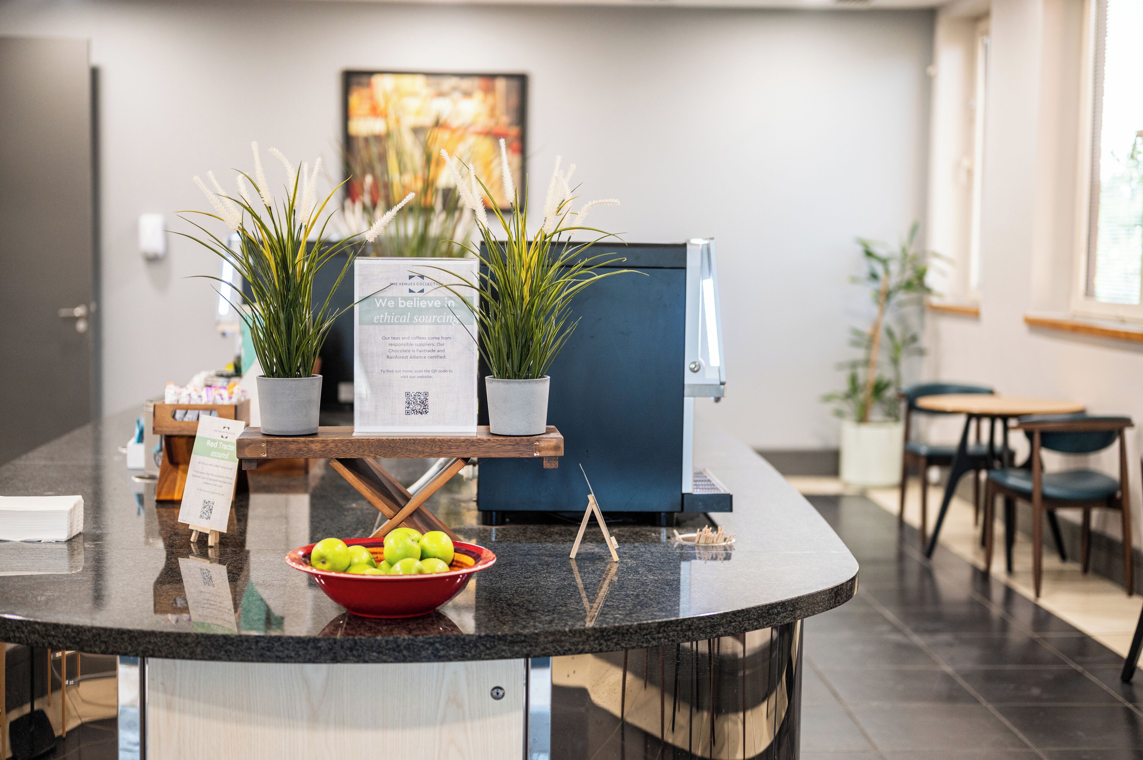 A reception area with a counter, two potted plants, a bowl of apples, and a sign about ethical sourcing.
