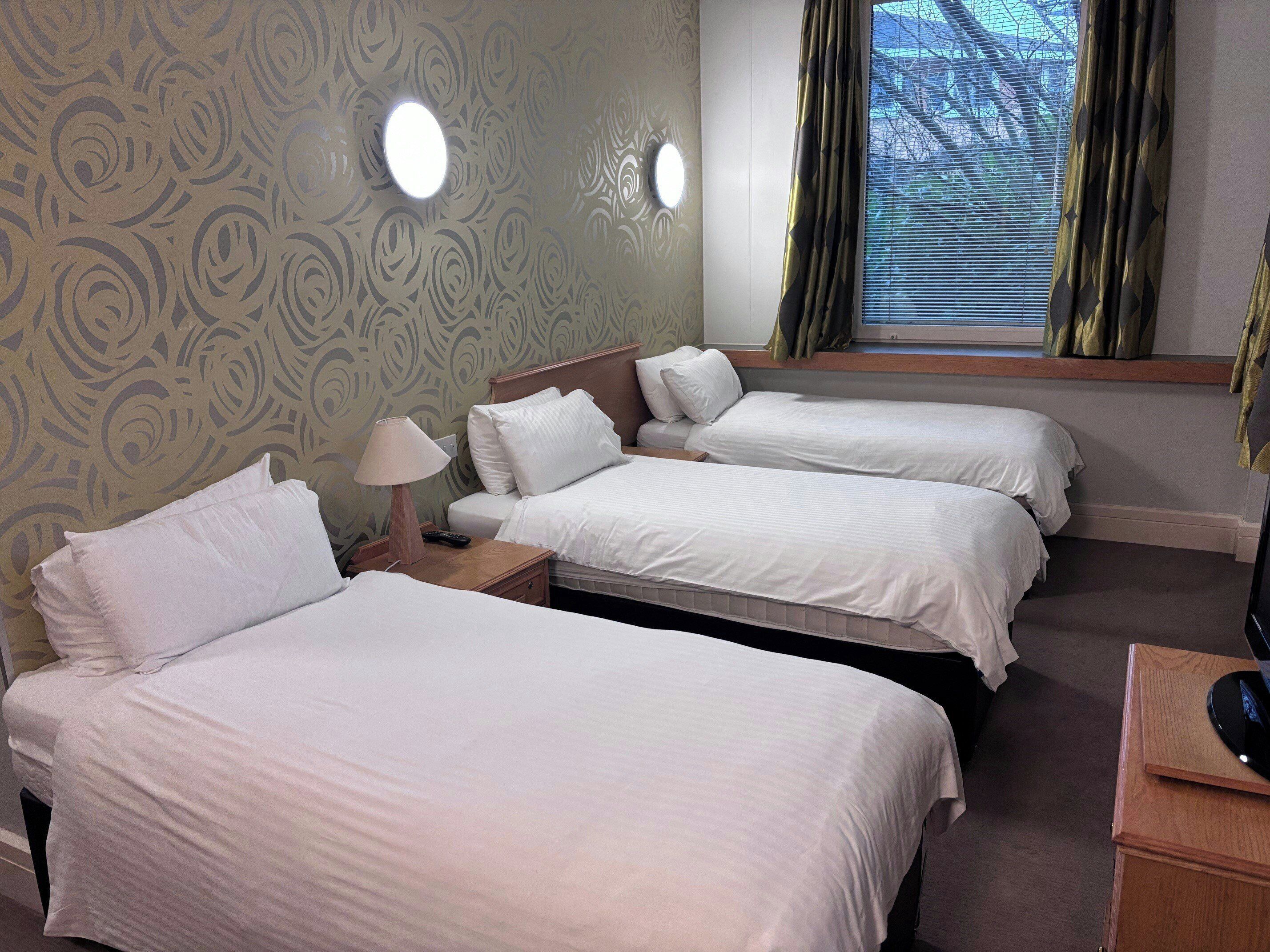 A hotel room with three single beds, a nightstand with a lamp, and a window with curtains.