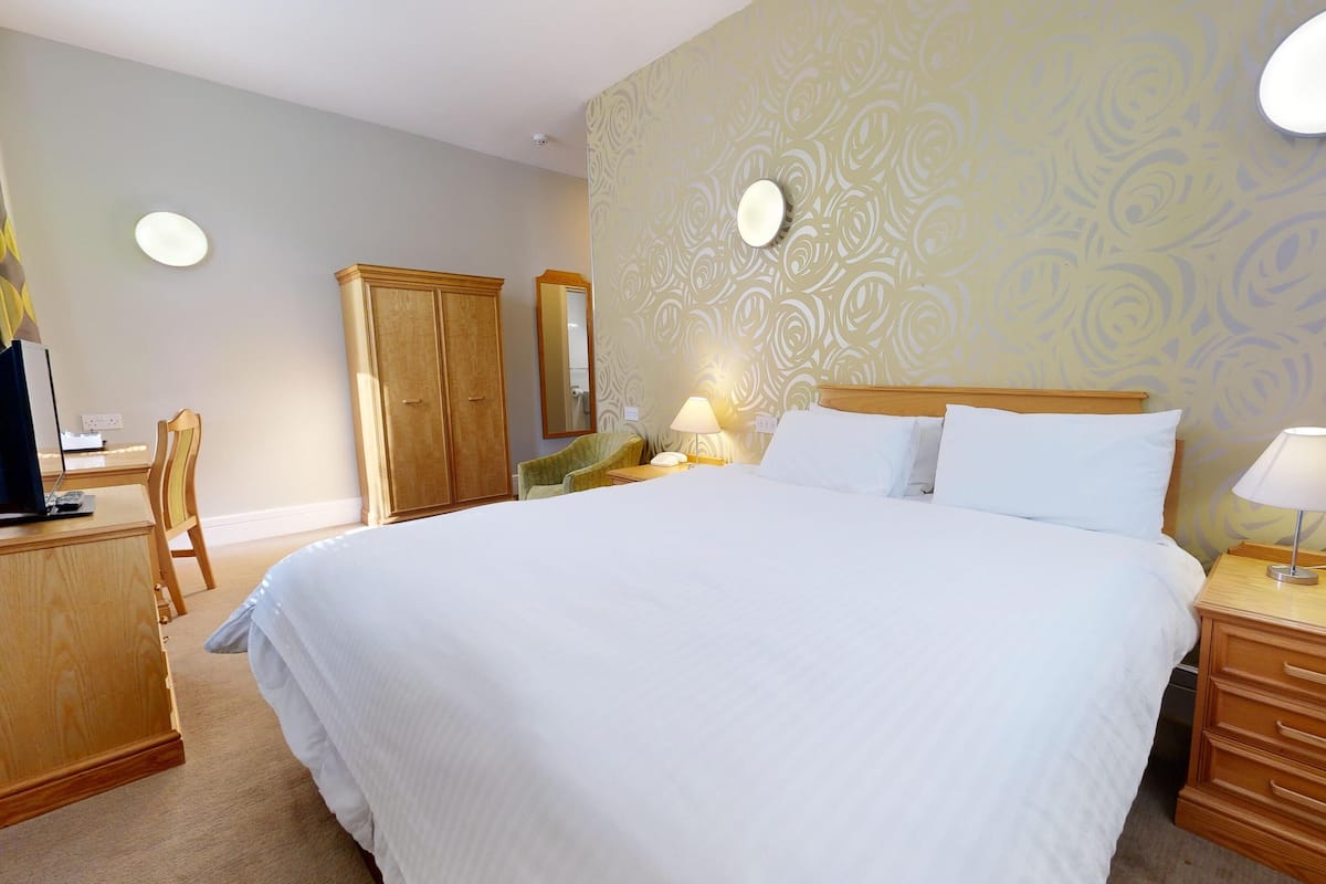 Une chambre d’hôtel avec un grand lit, une armoire en bois, un bureau avec une chaise, une table de chevet avec une lampe et un miroir au mur.