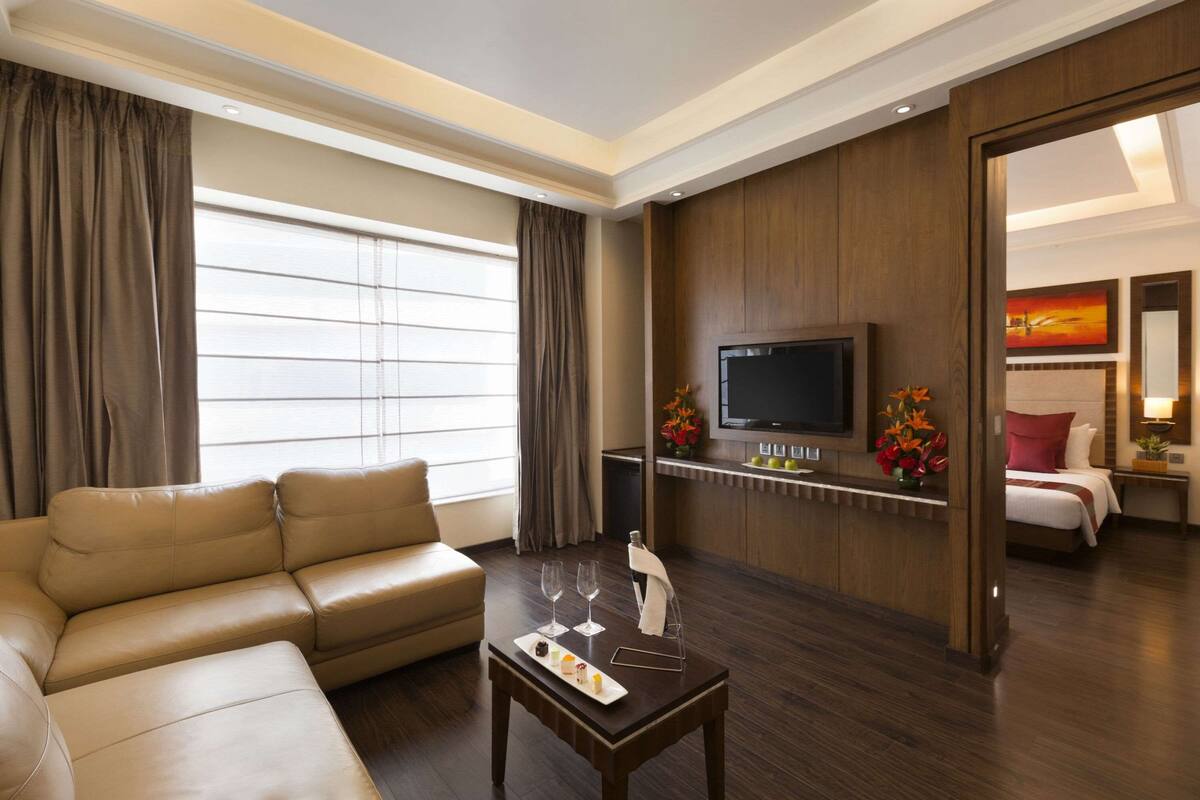 A hotel room with a leather sofa, a small table with a tray of snacks, a flat-screen TV mounted on the wall, and a bed with a headboard.