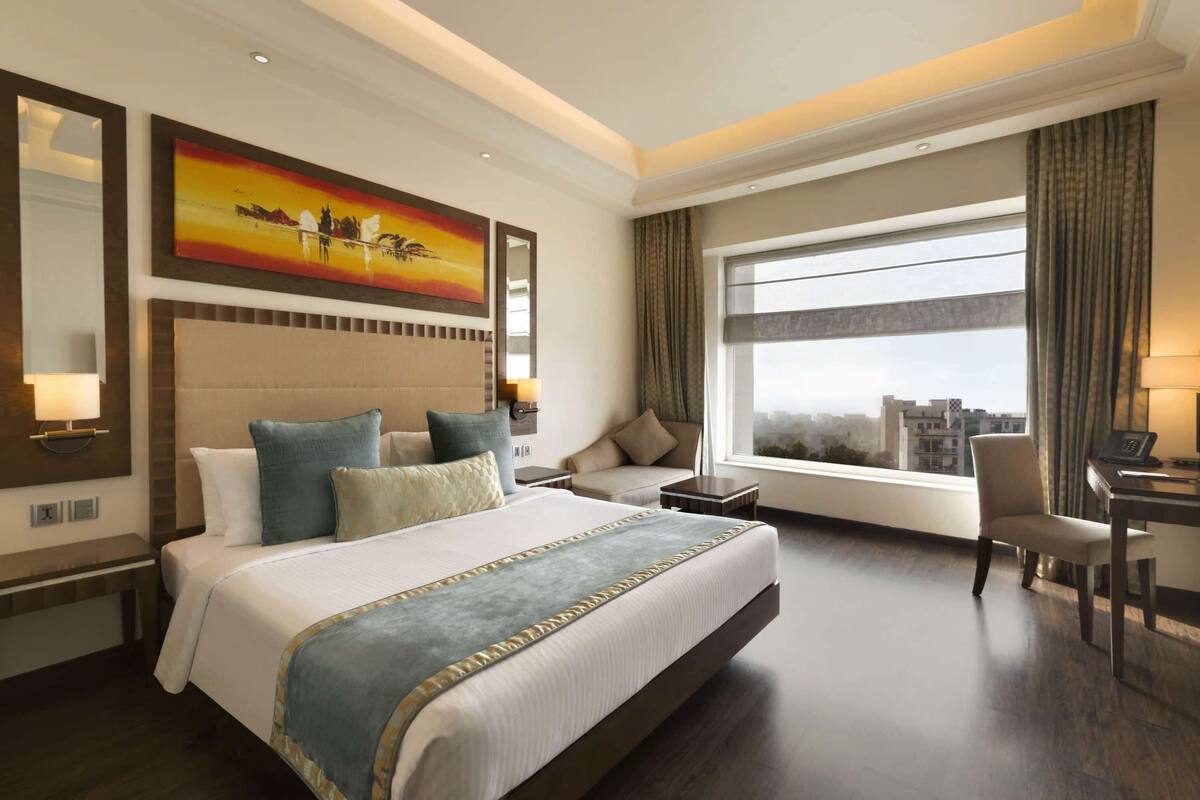 A hotel room with a large bed, a desk, a chair, a window with a city view, and a painting on the wall.
