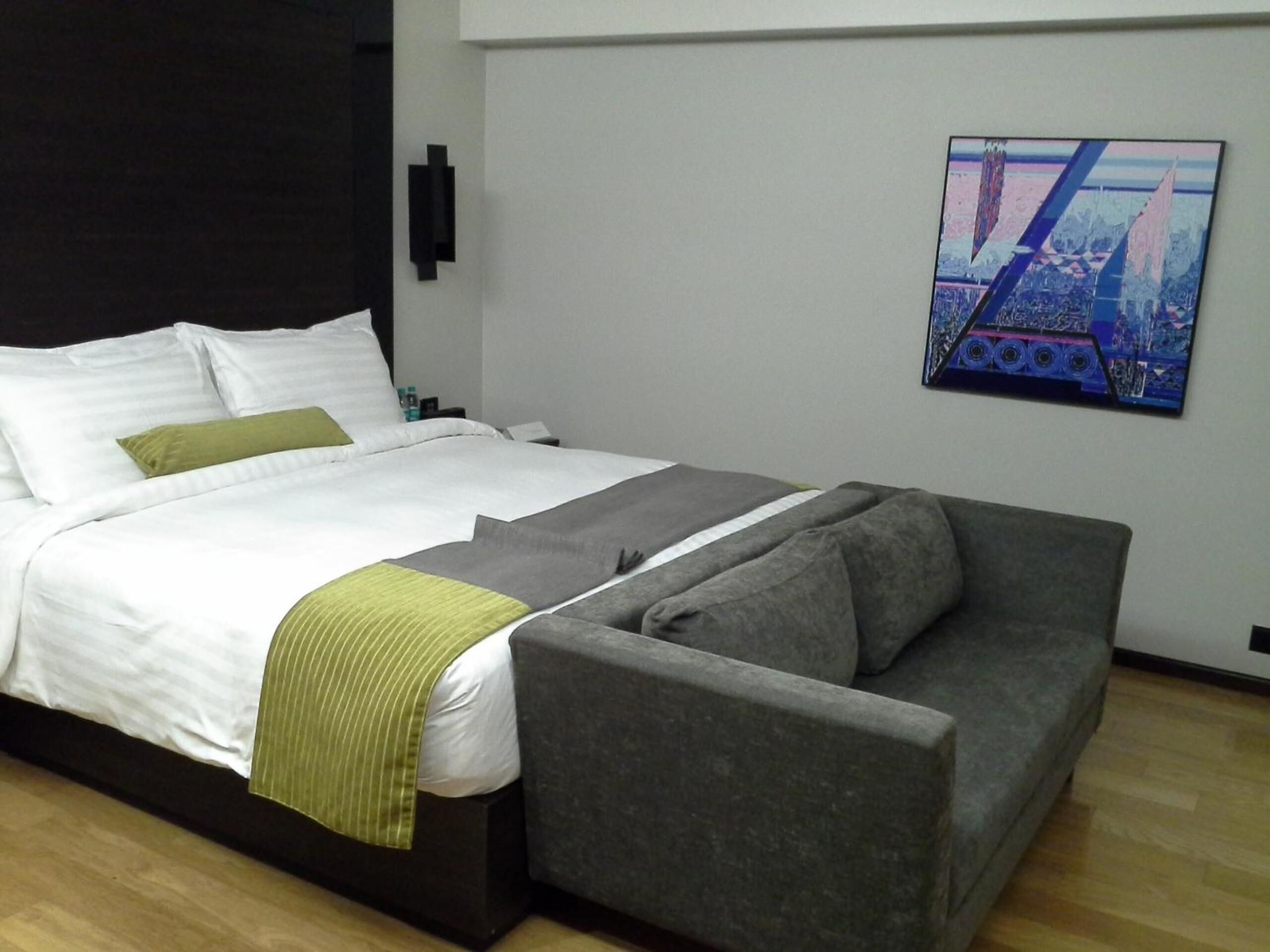 A modern hotel room with a large bed, grey and green bedding, a grey sofa, and a cityscape painting on the wall.