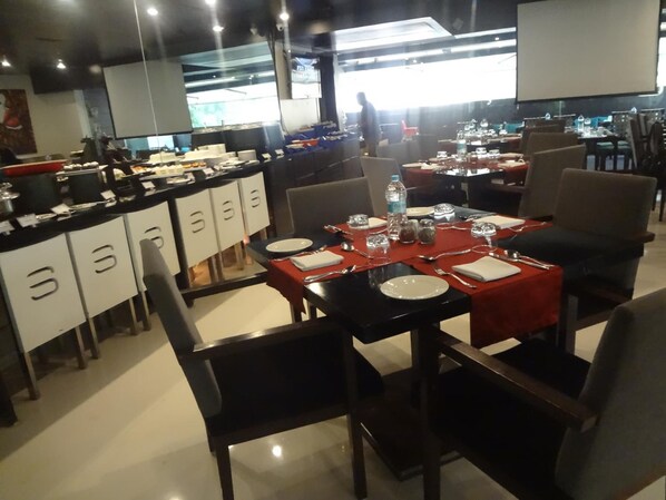 A restaurant with multiple tables and chairs, a counter with various items, and a large screen in the background. - Svenska Design Hotel, Mumbai (Mumbai)