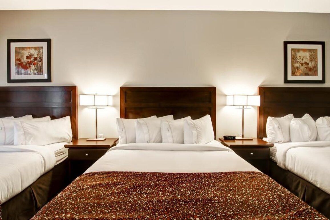 A hotel room with two beds, a large bed in the center and two single beds on either side. There are two bedside tables with lamps, and artwork is displayed on the walls.