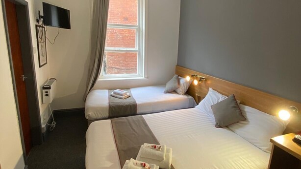 A hotel room with two beds, a TV mounted on the wall, a window with curtains, and a bedside table with a lamp. - Alumhurst Hotel (Blackpool)