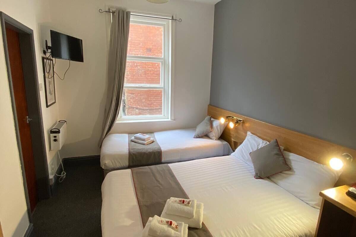 A hotel room with two beds, a TV mounted on the wall, a window with curtains, and a bedside table with a lamp.