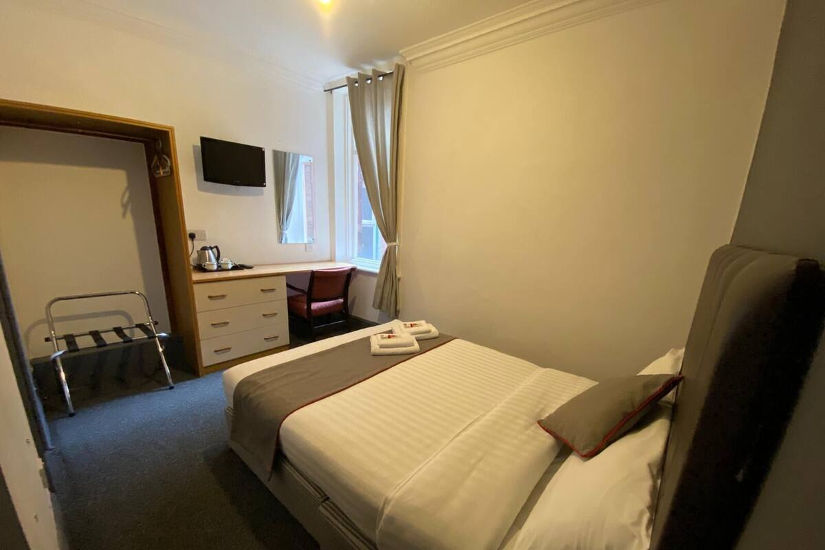 A hotel room with a bed, a desk with a chair, a TV mounted on the wall, and a window with curtains.
