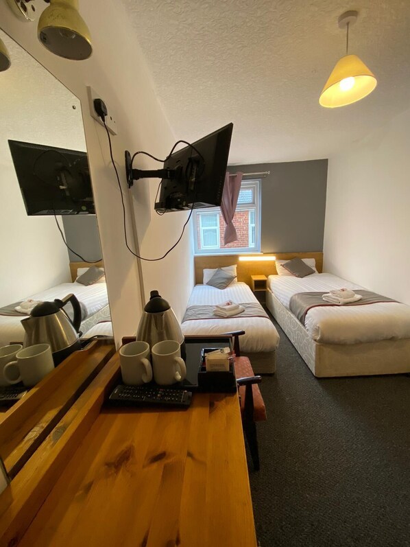 A hotel room with two single beds, a desk with a computer and coffee maker, a TV mounted on the wall, and a window with a view of a building. - Alumhurst Hotel (Blackpool)