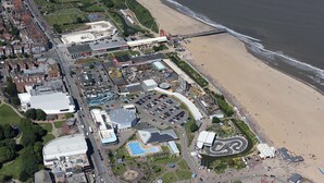 An aerial view of a coastal area with a beach, a roller coaster, and various buildings including a large stadium and parking areas. - Alumhurst Hotel (Blackpool)