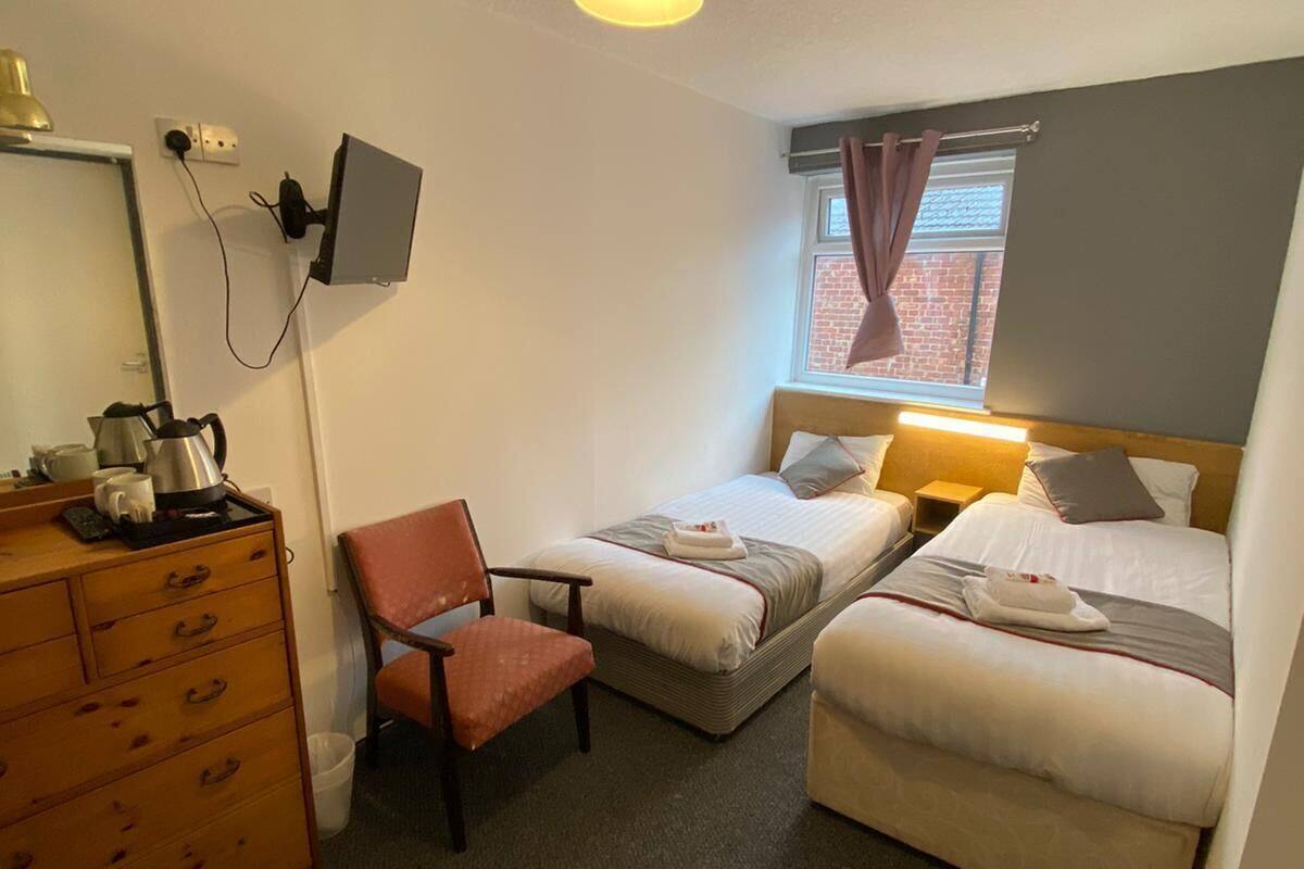 A small, tidy room with two single beds, a wooden dresser, a chair, a desk with a computer monitor, and a window with curtains.