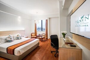 Quarto de hotel com cama, escrivaninha, cadeira e televisão exibindo o nome do hotel.