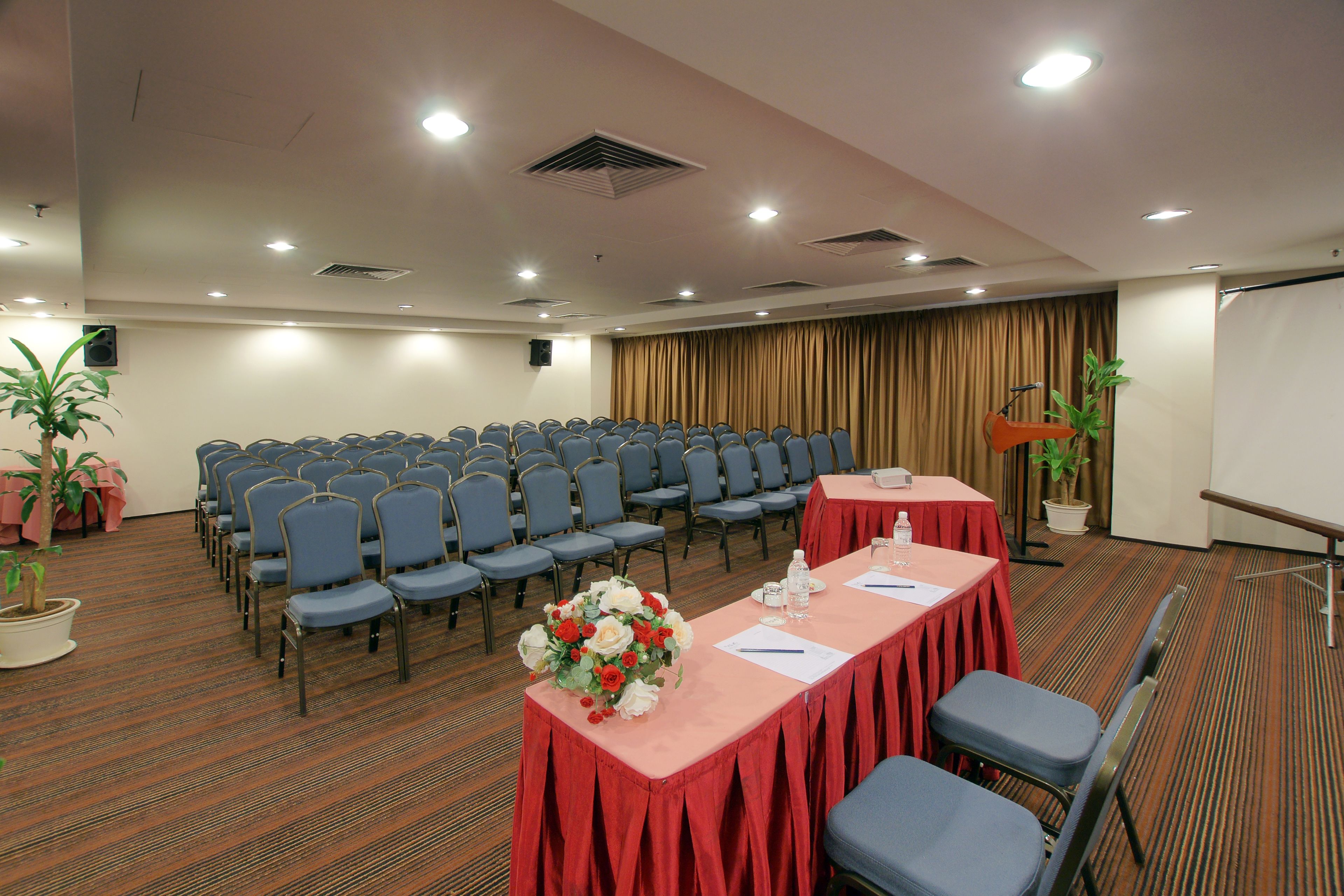 Una sala conferenze con file di sedie, un tavolo con tovaglia rossa e uno schermo per proiettore.