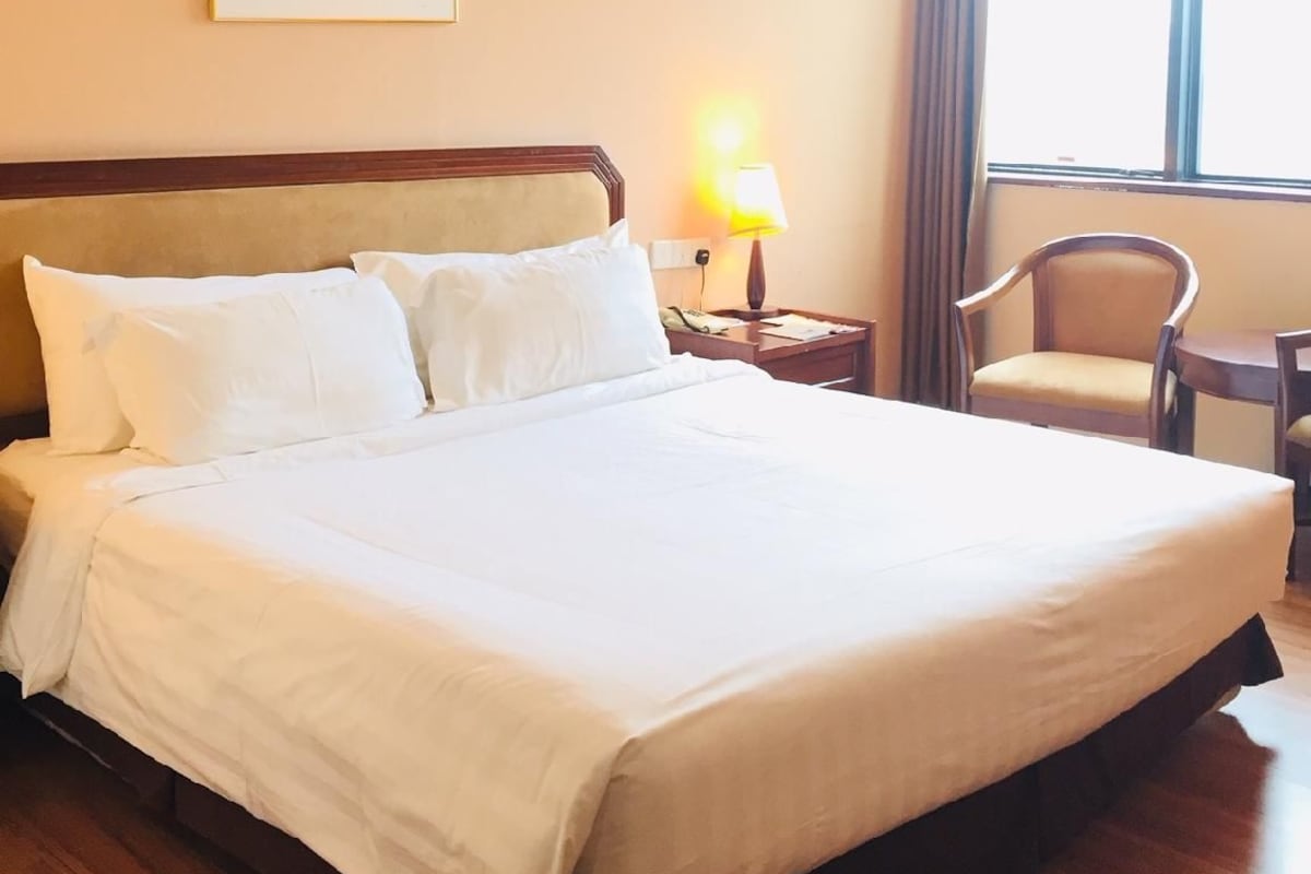 Una habitación de hotel con una cama grande, una lámpara de noche, una silla y un ventanal con cortinas.