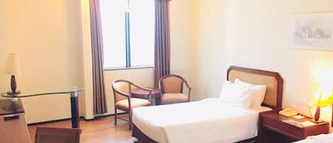 A hotel room with a bed, a chair, a desk, and a nightstand with a lamp.