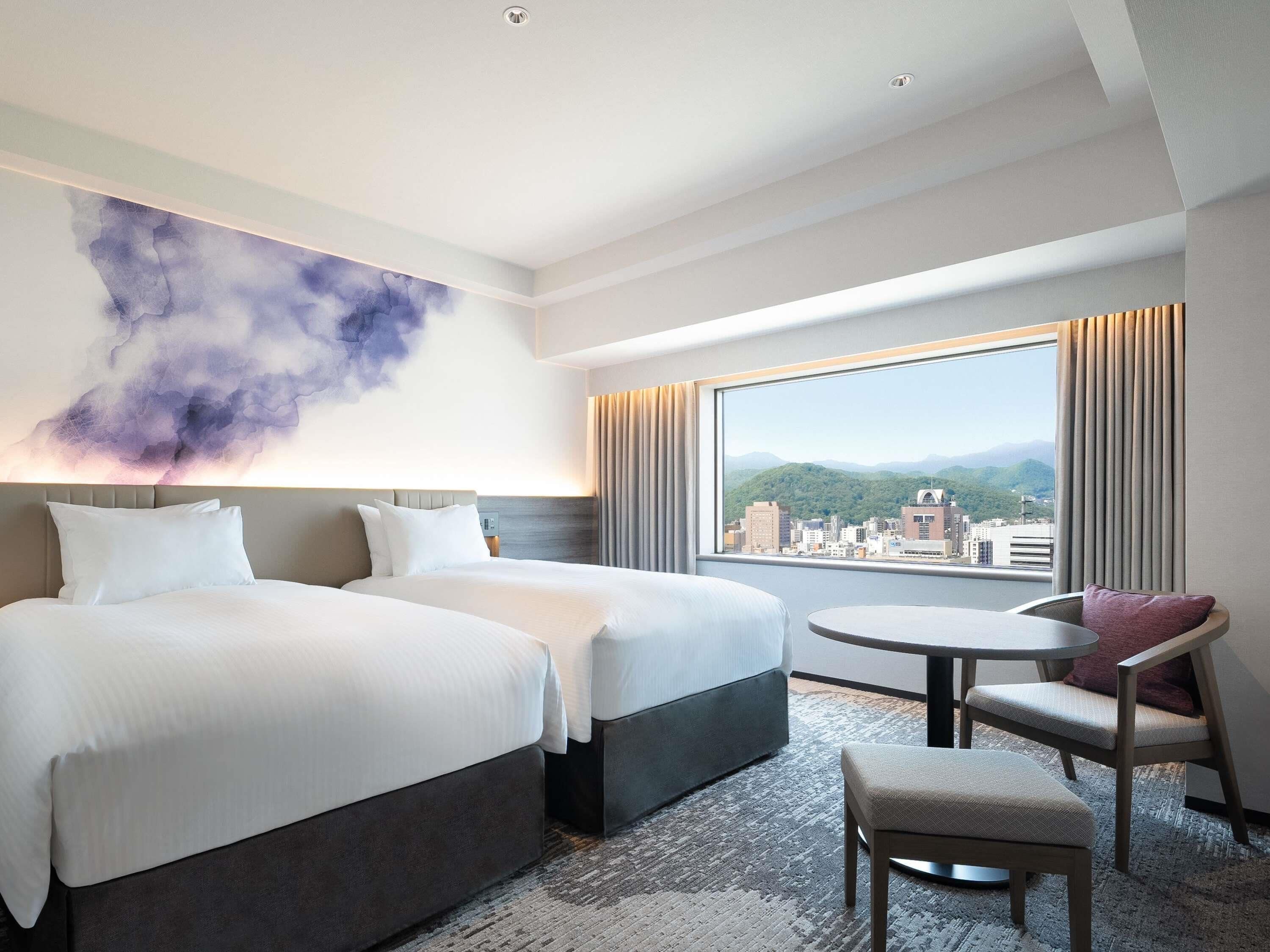 A hotel room with two beds, a round table, a chair, a large window with a city view, and a wall art piece.