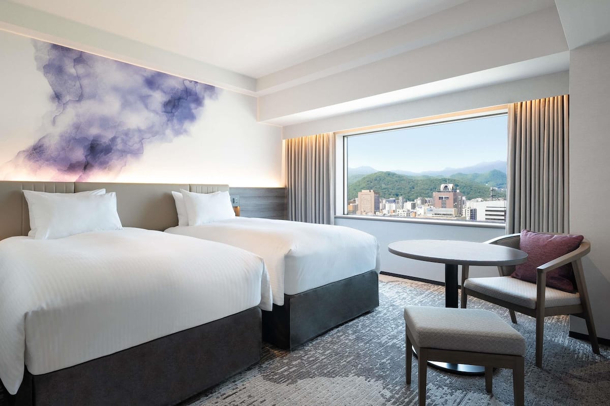 A hotel room with two beds, a round table, a chair, a large window with a city view, and a wall art piece.