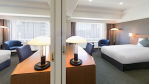 A hotel room with a bed, a desk with a lamp, a chair, and a view of the city through large windows.