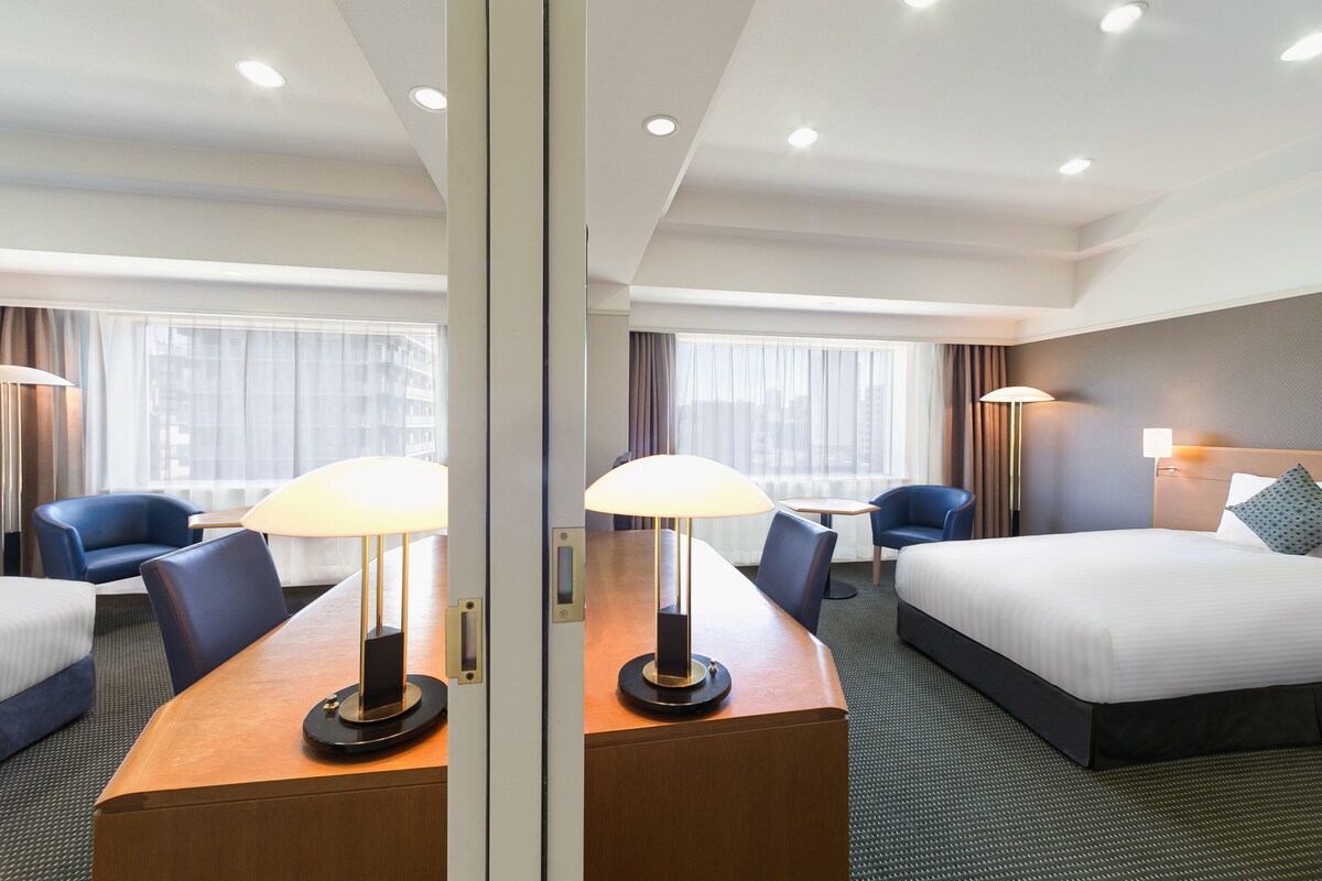 A hotel room with a bed, a desk with a lamp, a chair, and a view of the city through large windows.