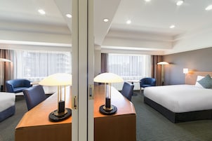 A hotel room with a bed, a desk with a lamp, a chair, and a view of the city through large windows.