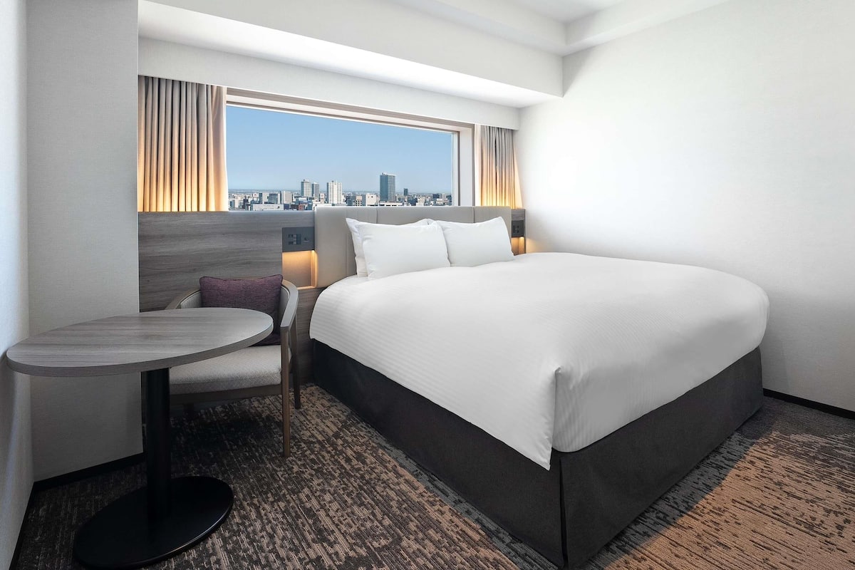 Quarto de hotel com cama, uma mesinha, uma cadeira, janela com vista para a cidade e paisagem urbana visível através da janela.