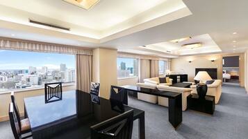 A modern hotel room with a dining area, a sofa set, a desk, and a view of the cityscape.
