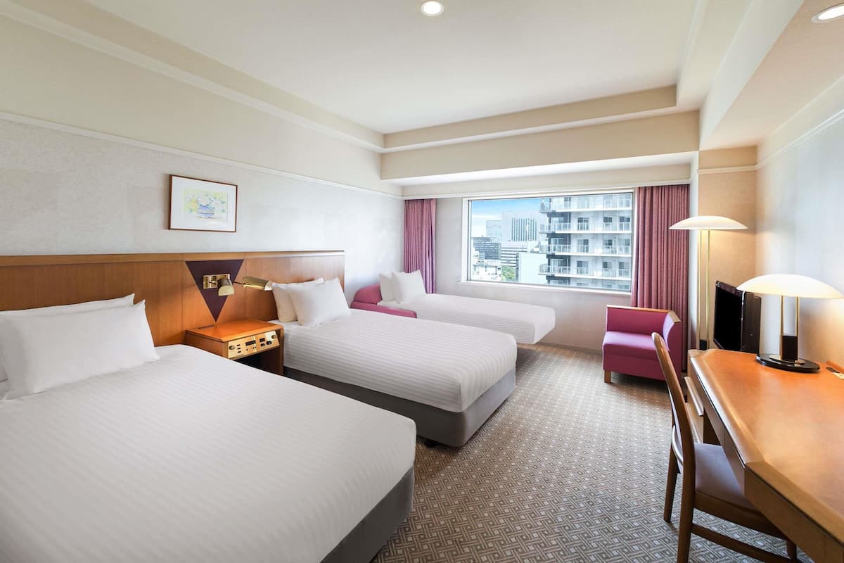 Quarto de hotel com duas camas, uma escrivaninha, uma cadeira, uma televisão e uma janela com vista para a cidade.
