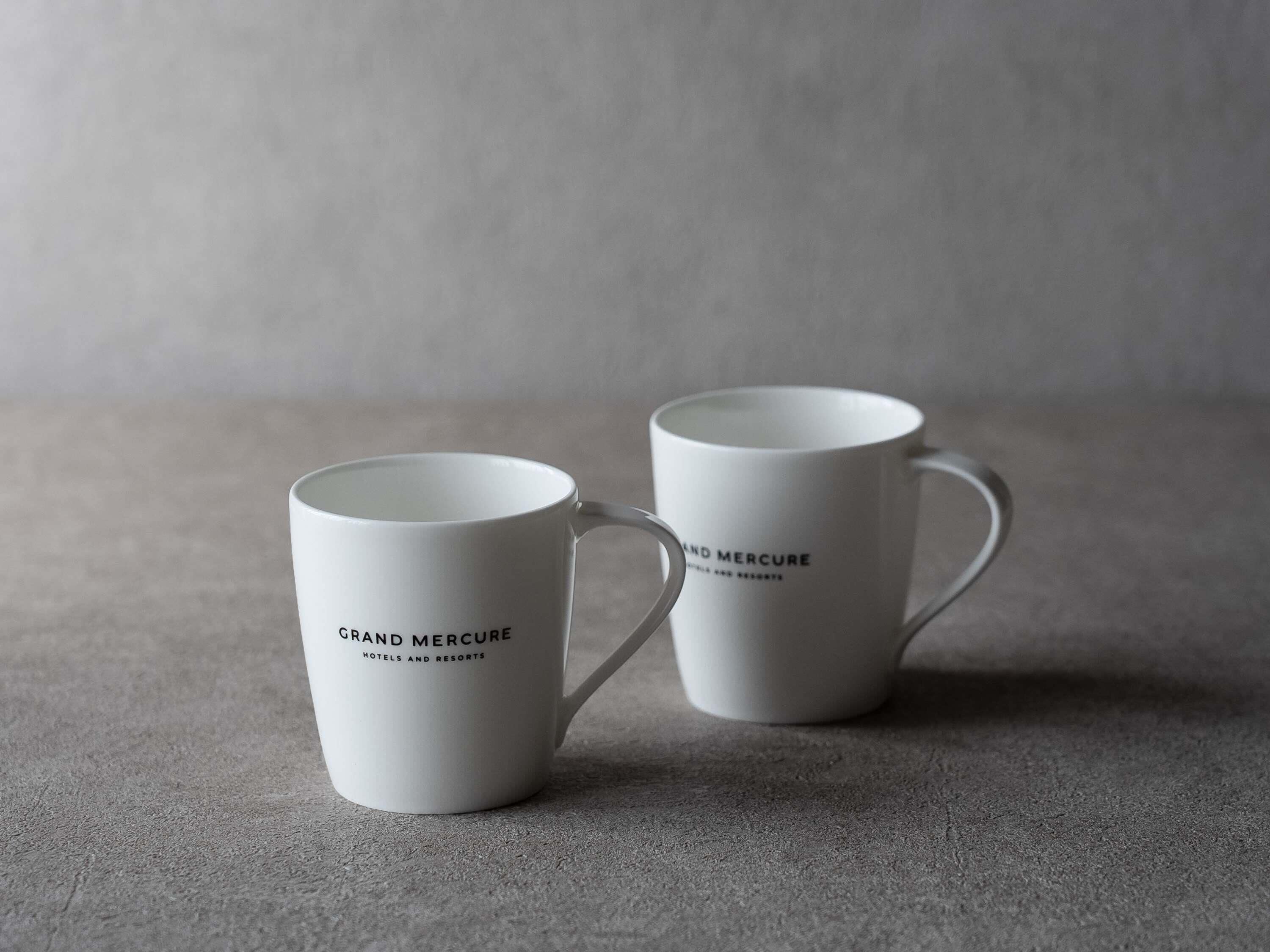Deux tasses blanches ornées du texte « Grand Mercure Hotels and Resorts », posées sur une surface texturée.