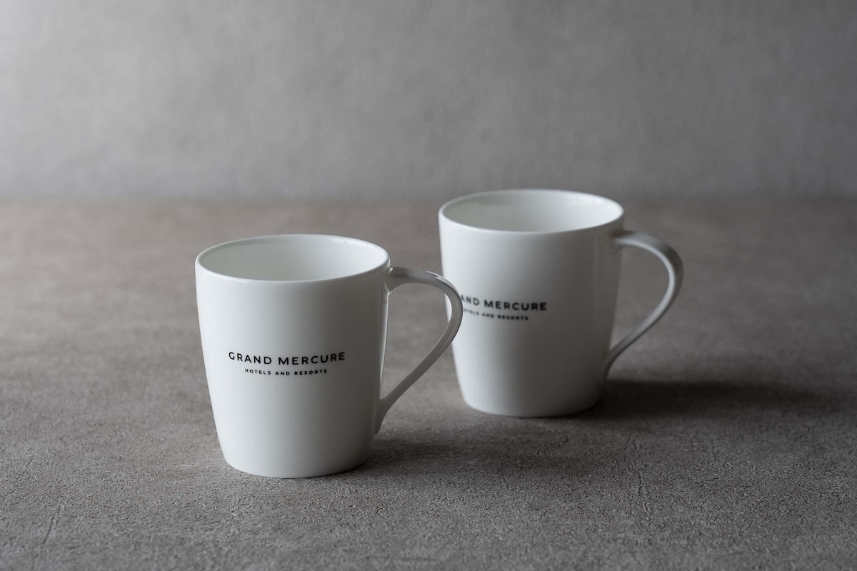 Deux tasses blanches ornées du texte « Grand Mercure Hotels and Resorts », posées sur une surface texturée.
