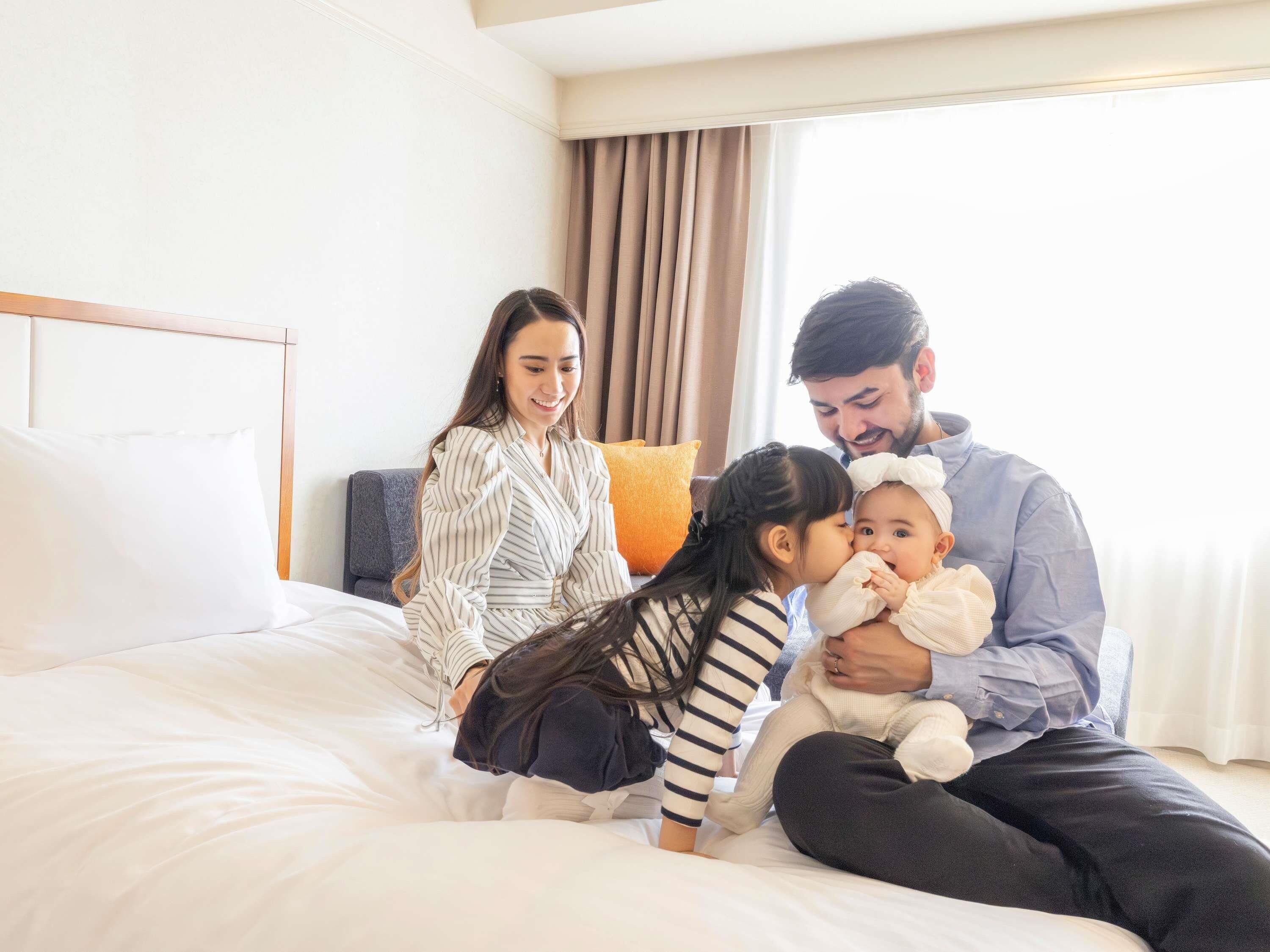 Una familia de cuatro personas en una cama con cabecera, una ventana con cortinas y una almohada.