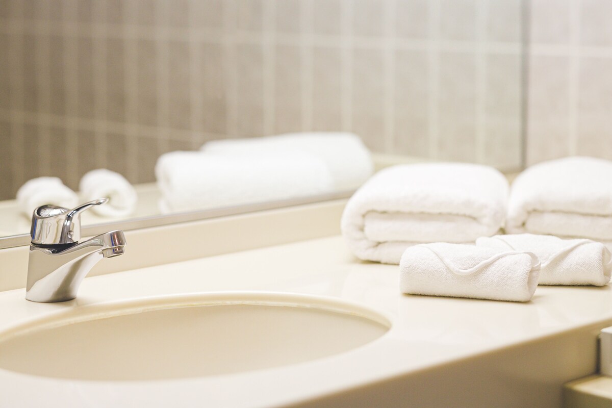 A bathroom sink with a chrome faucet and a mirror above it. There are several neatly folded white towels stacked on the counter.