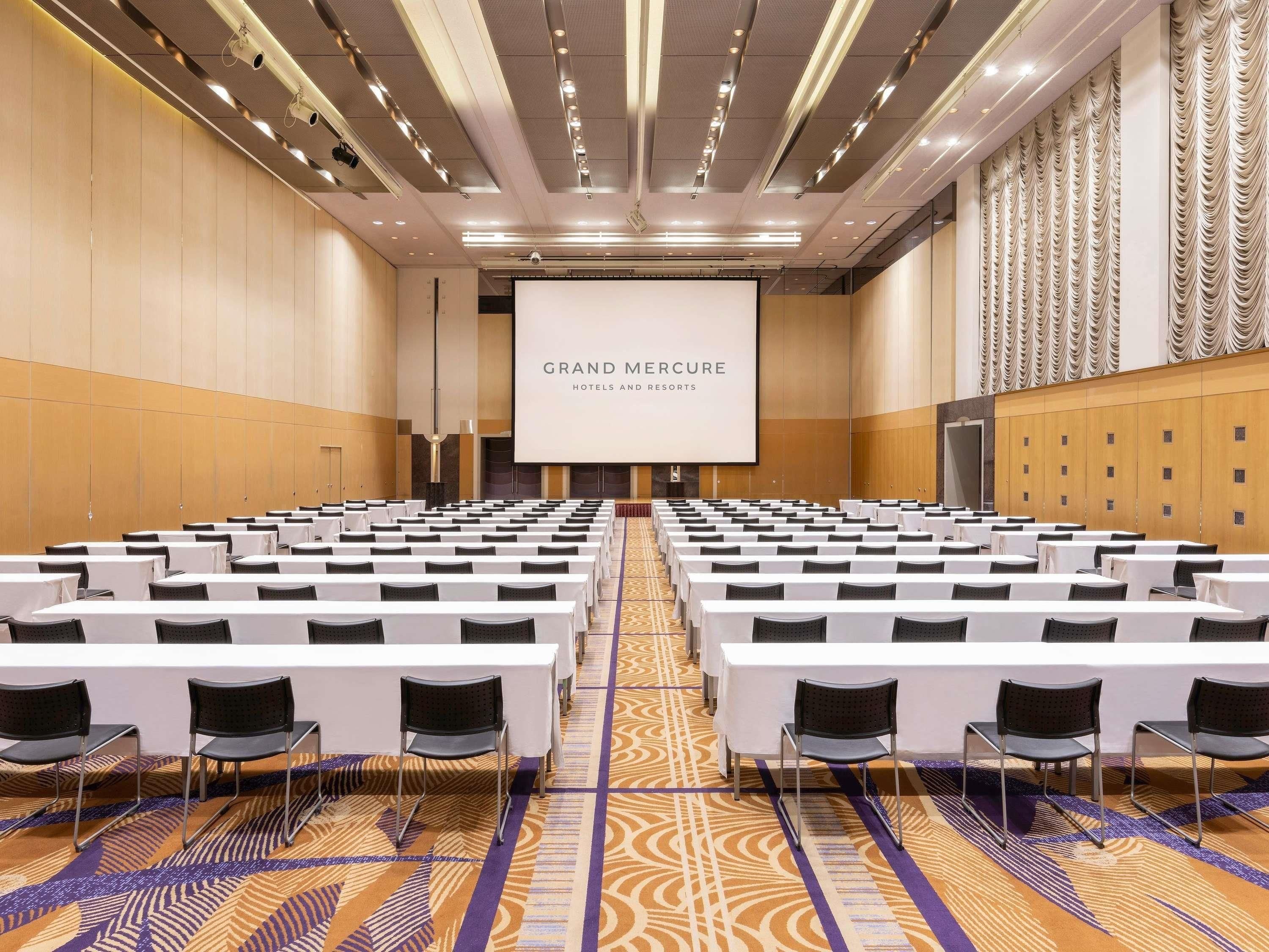A conference room with rows of tables and chairs, a large screen displaying 'GRAND MERCURE HOTELS AND RESORTS', and a patterned carpet.