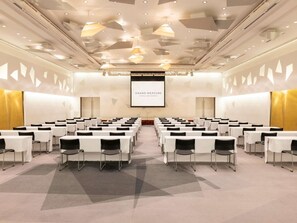 Un salón de conferencias con filas de mesas y sillas, una pantalla grande que muestra 'GRAND MERCURE HOTELS AND RESORTS' y un diseño geométrico en el techo.