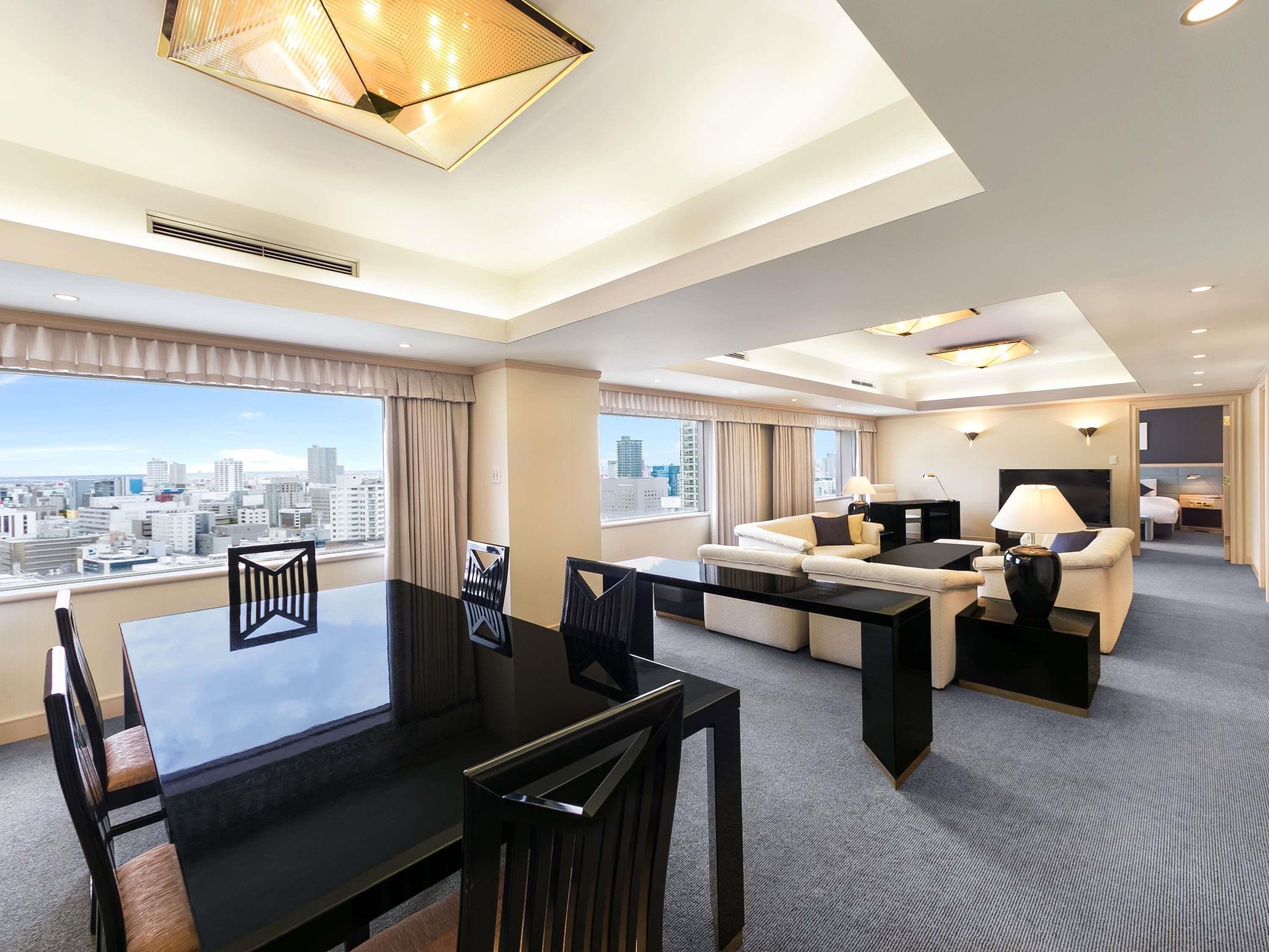 A modern hotel room with a dining area, a sofa set, a desk, and a city view.