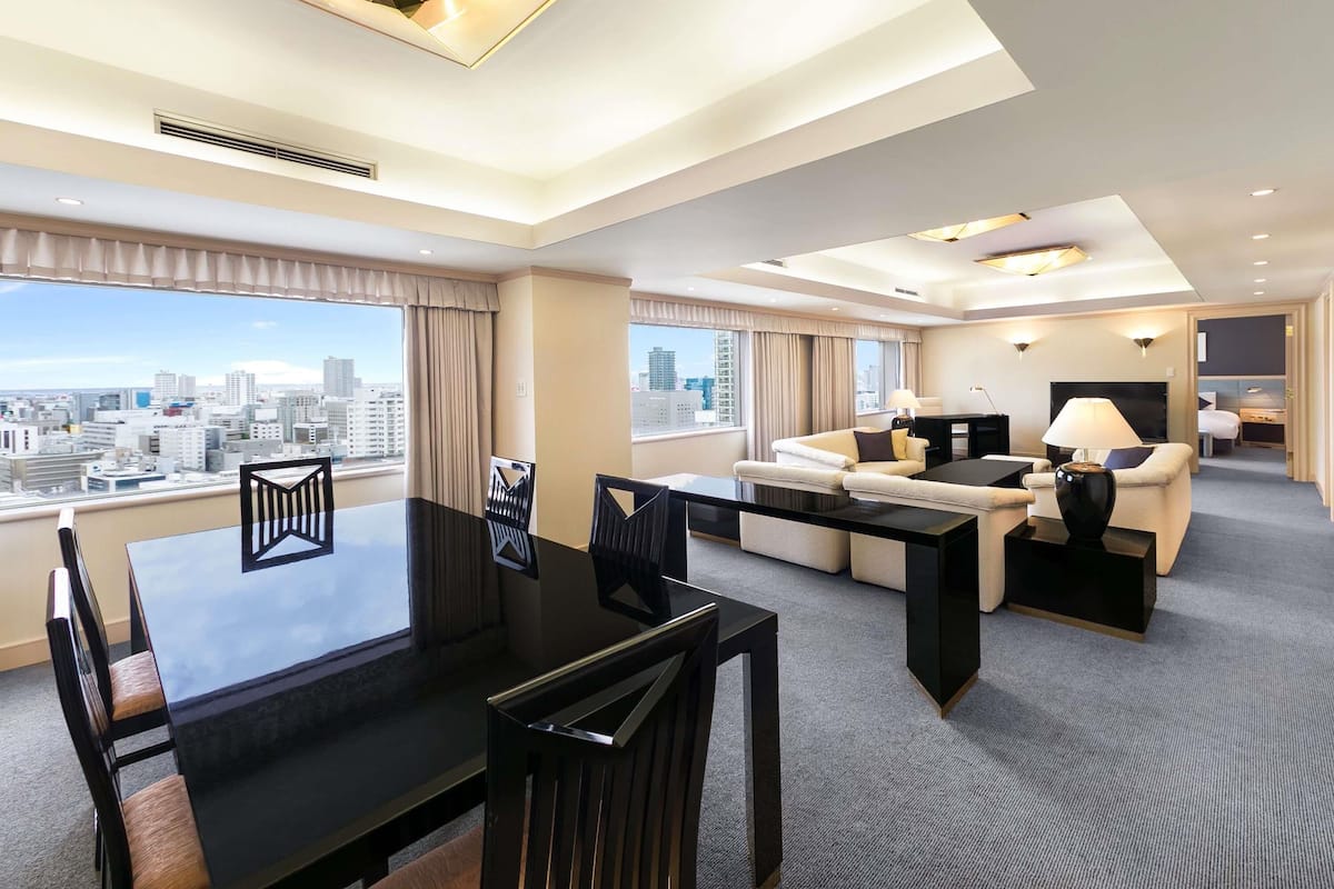 A modern hotel room with a dining area, a sofa set, a desk, and a city view.