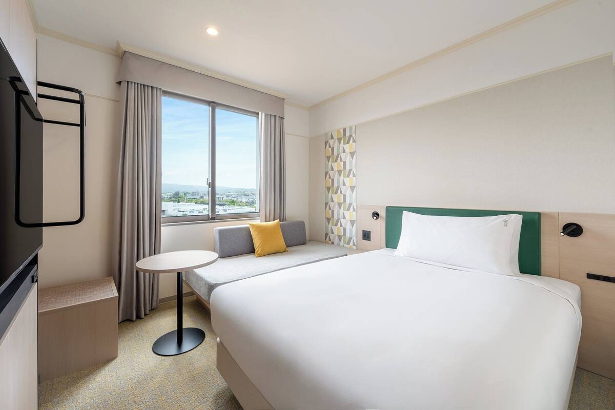 Une chambre d’hôtel avec un lit, une petite table ronde, une télévision et une fenêtre avec des rideaux.