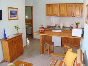 A compact kitchen with wooden cabinets, a small refrigerator, and a dining area with a chair and a table. - Bungalows Castillo Playa (Antigua)