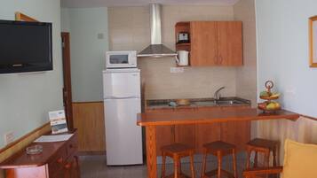 Una cocina compacta con refrigerador, microondas y armarios de madera. Se observa una pequeña isla con taburetes y una exhibición de frutas.