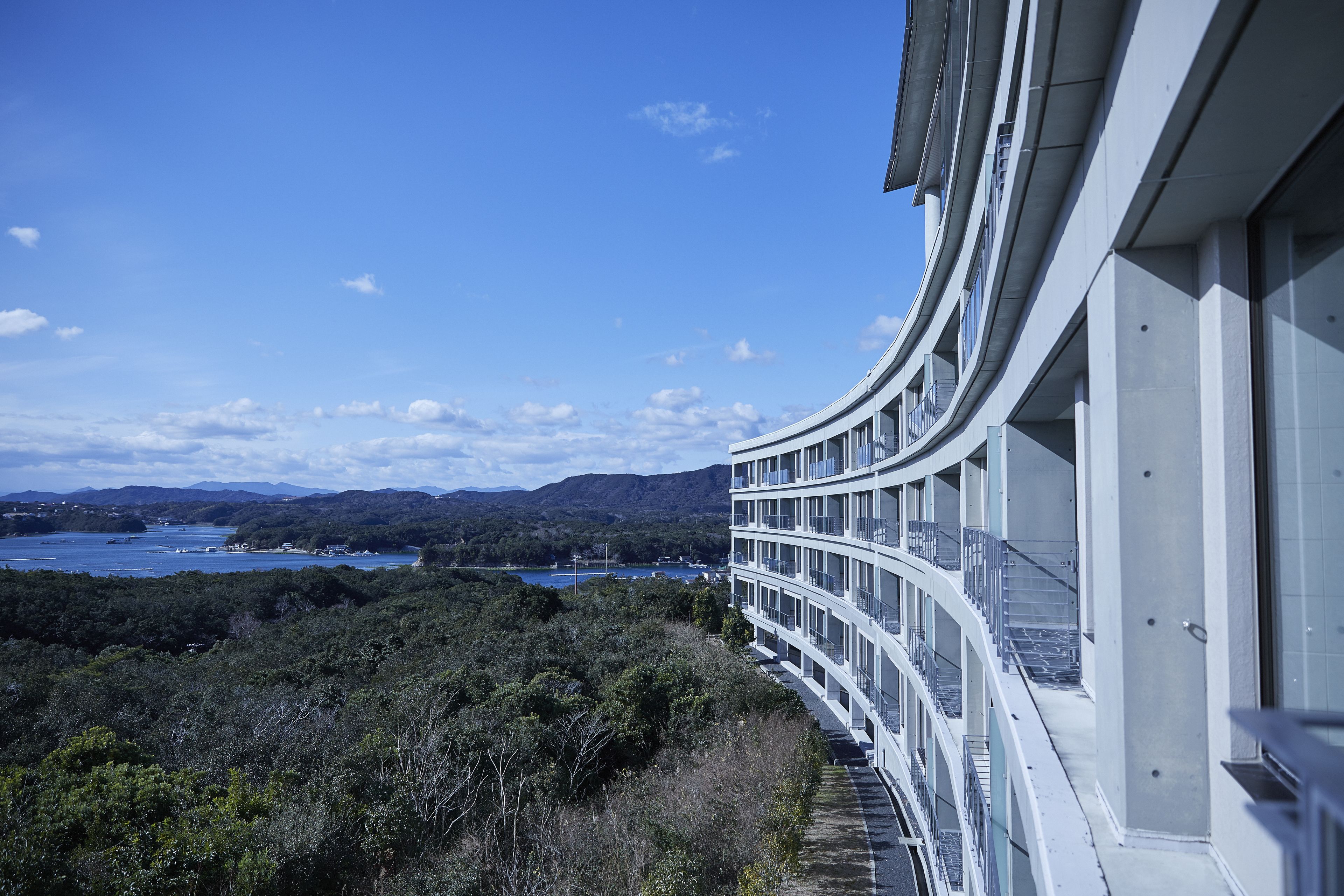 A modern building with a curved balcony overlooking a coastal landscape with mountains and a body of water.